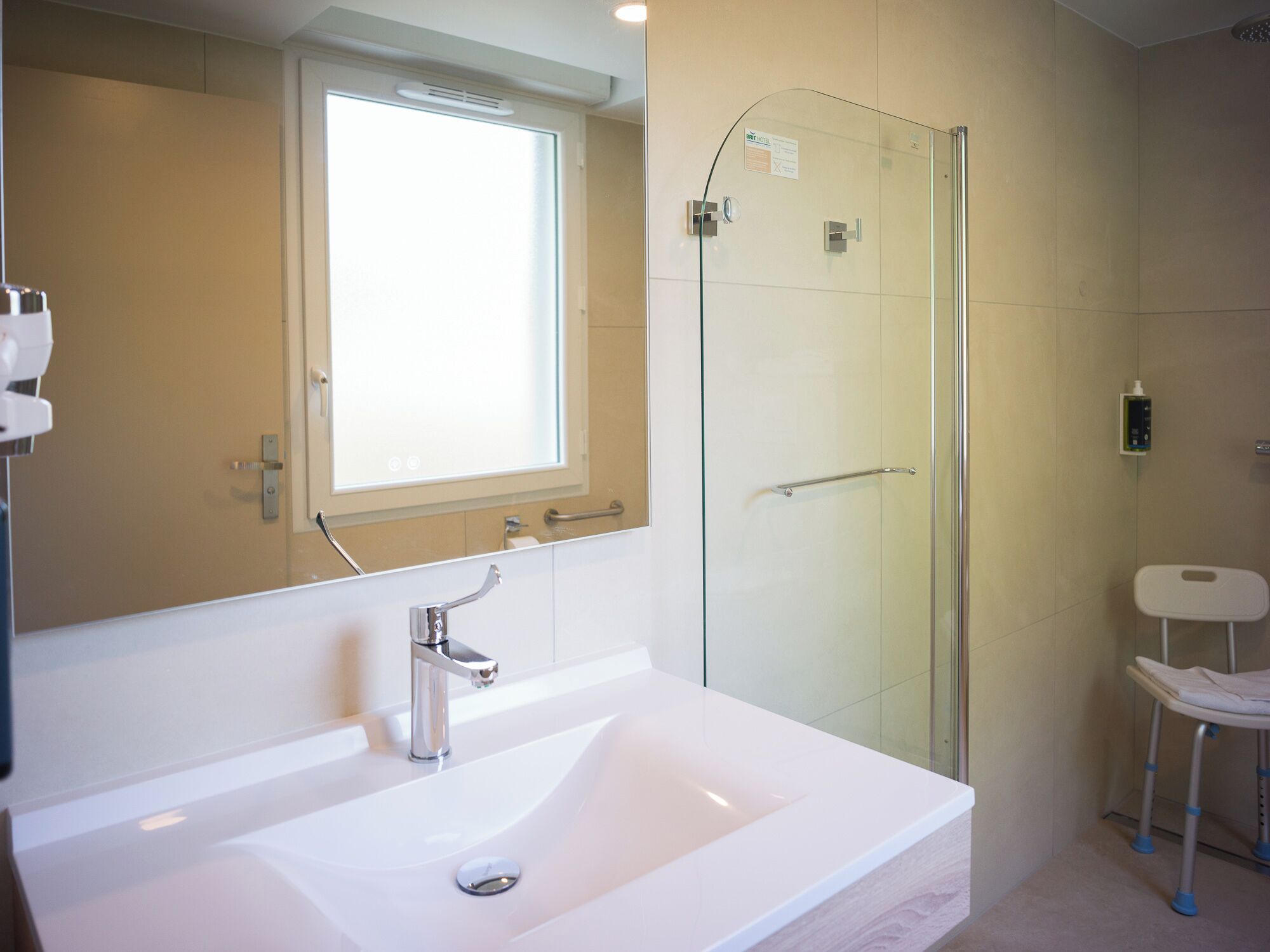 A modern bathroom with a large white sink, a glass shower enclosure, and a window above the mirror.