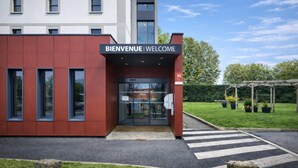 A modern building with a 'BIENVENUE | WELCOME' sign, a glass entrance, and a crosswalk leading to a parking area. - BRIT HOTEL CONFORT ROISSY SAINT-WITZ (Saint-Witz)