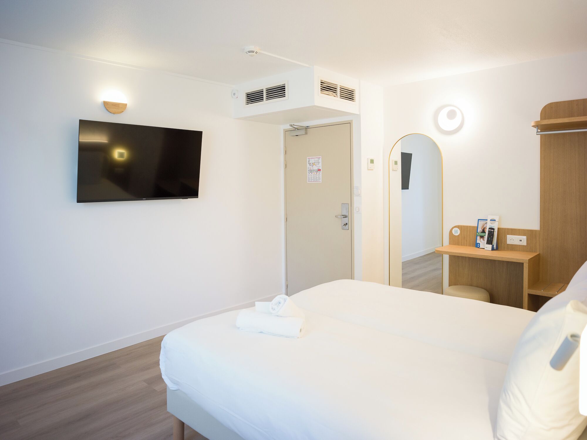 A hotel room with a bed, a flat-screen TV mounted on the wall, a wooden bedside table with a remote control, a mirror, and a door leading to another room.