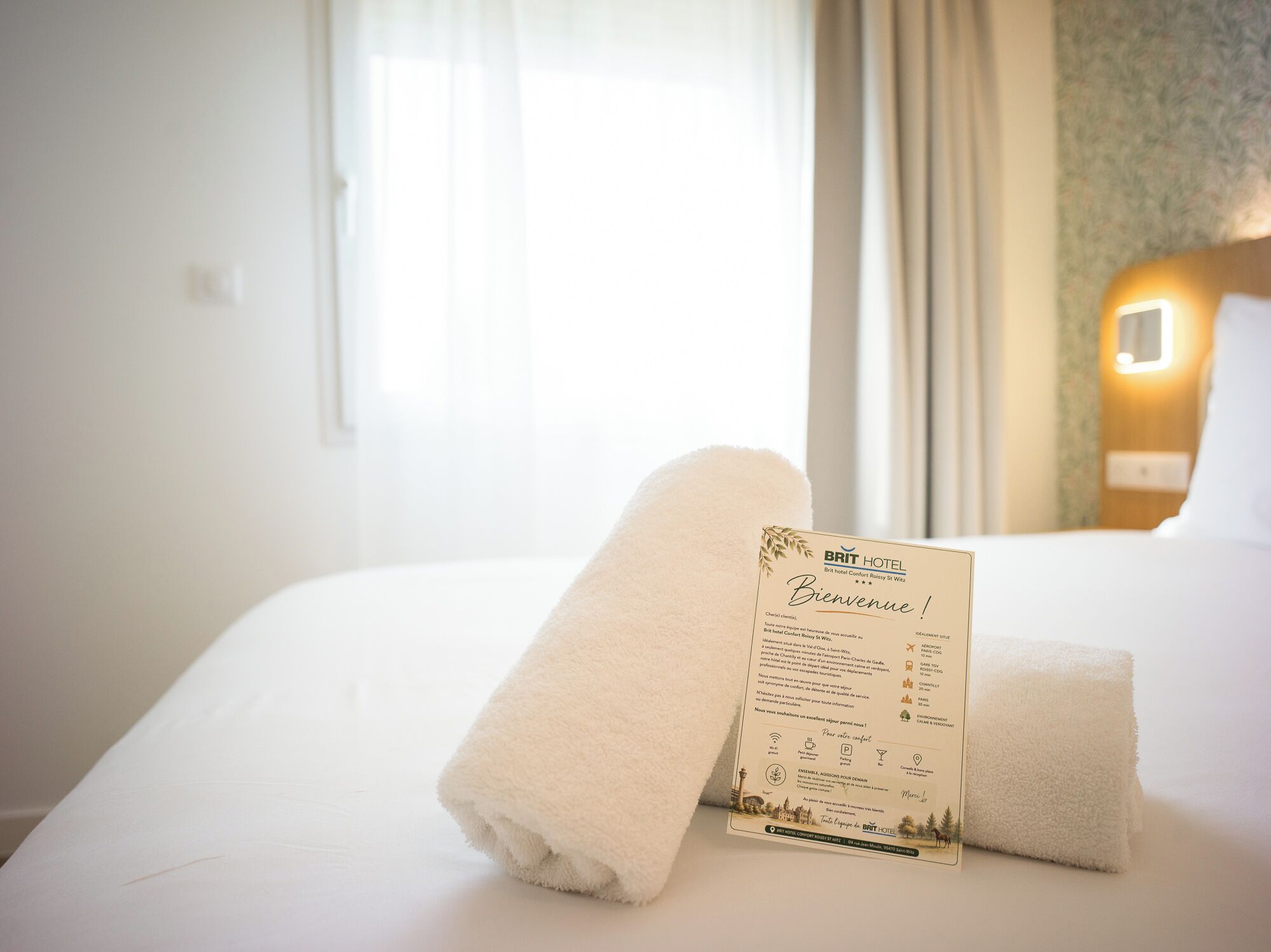 Een hotelkamer met een net opgemaakt bed, een welkomstbord en een raam met gordijnen.