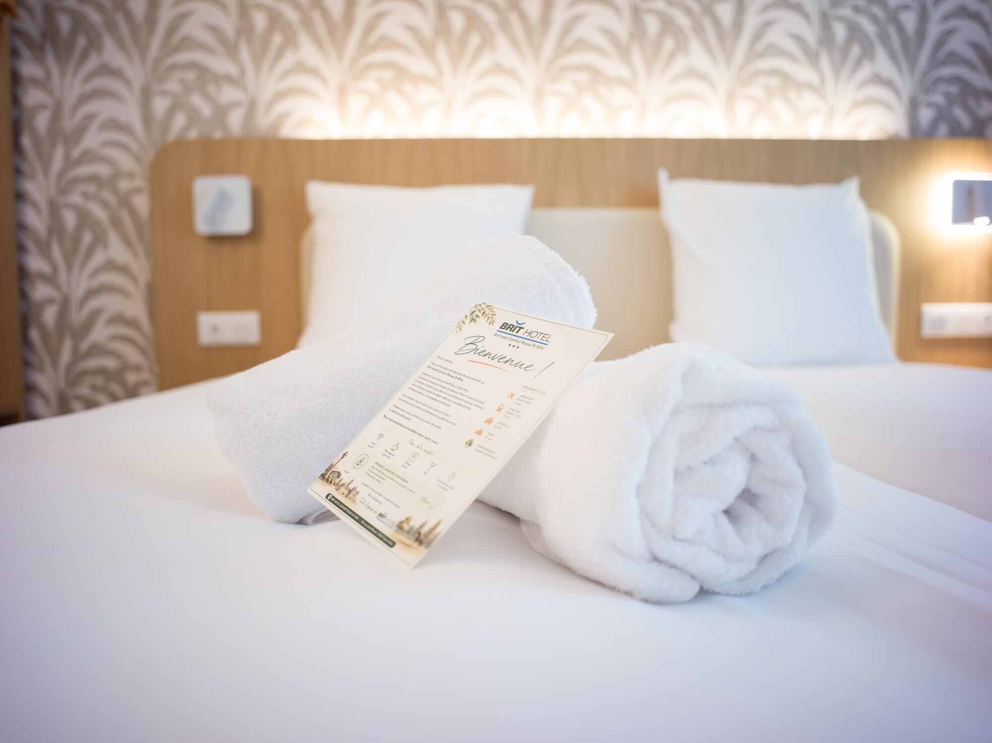 Una cama bien tendida con una toalla enrollada y una tarjeta de bienvenida impresa, sobre un fondo de papel tapiz estampado.