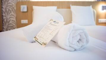 Una cama bien tendida con una toalla enrollada y una tarjeta de bienvenida impresa, sobre un fondo de papel tapiz estampado.