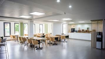 Un grand espace de salle à manger avec plusieurs tables et chaises, un coin cuisine équipé d’un réfrigérateur et d’une machine à café, et de grandes fenêtres laissant entrer la lumière naturelle.