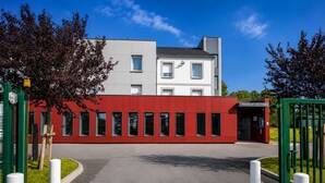 Un edificio moderno con fachada roja y sección superior gris, una entrada protegida por una reja y un cielo despejado.