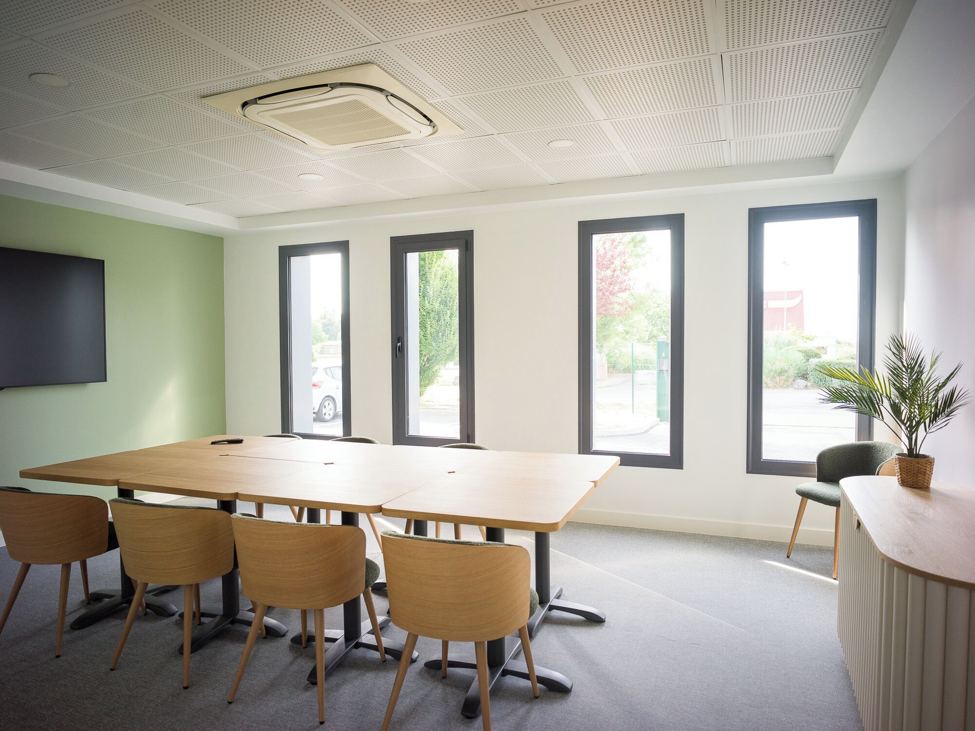 A conference room with a large table, four chairs, a TV on the wall, and three windows.