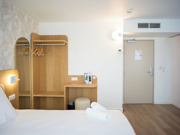 Habitación de hotel con cama, armario de madera, escritorio con silla, un taburete pequeño y una puerta.