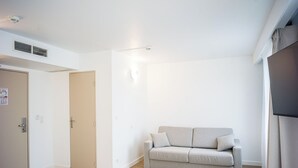 A minimalist room with a beige sofa, a white door, and a flat-screen TV on the wall. - BRIT HOTEL CONFORT ROISSY SAINT-WITZ (Saint-Witz)