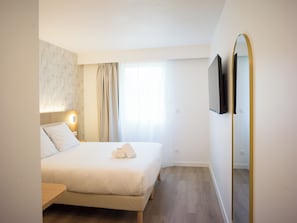 Une chambre d’hôtel avec un lit, une table de chevet, une lampe, un miroir et une fenêtre avec des rideaux.