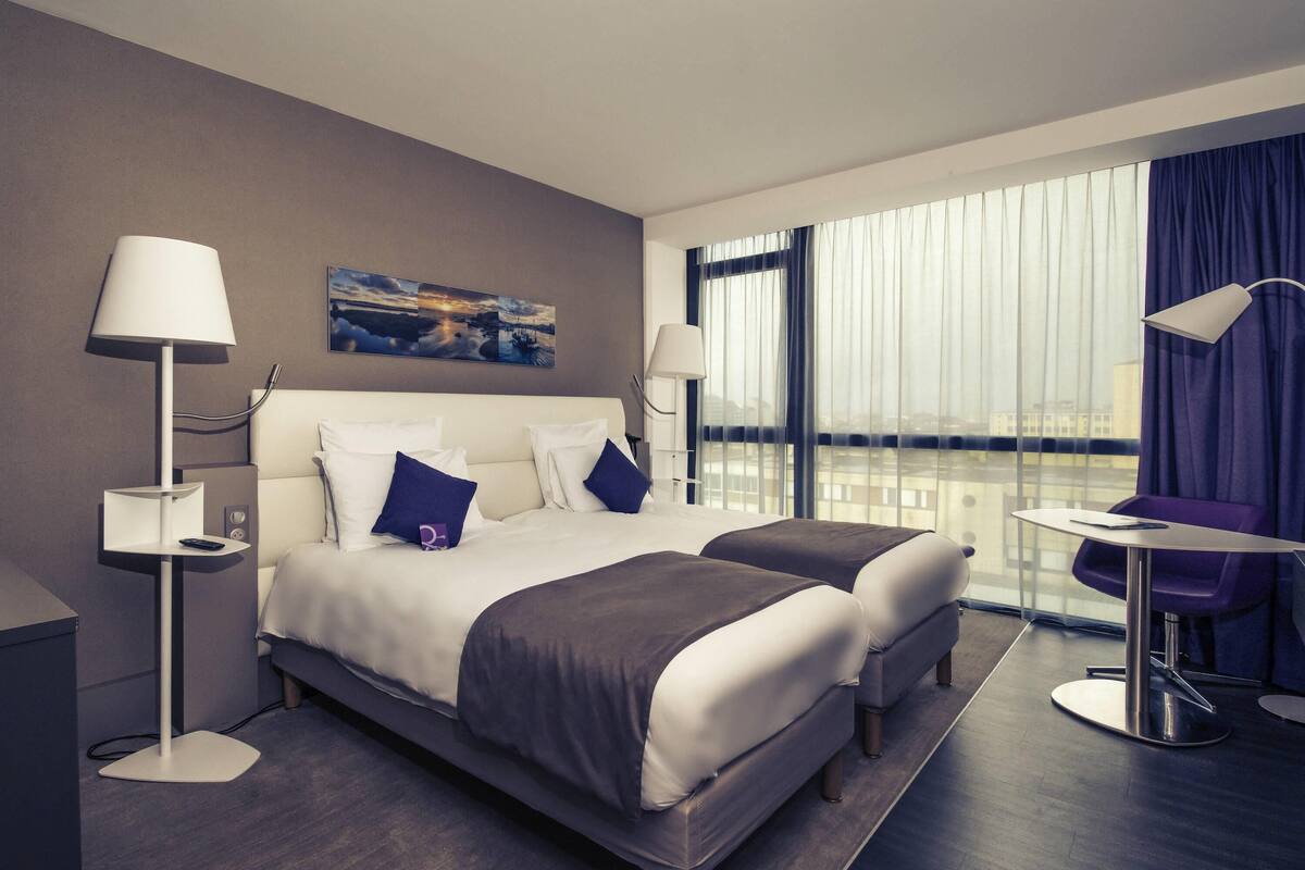 A hotel room with a bed, two bedside tables, a desk with a chair, and a large window with curtains.