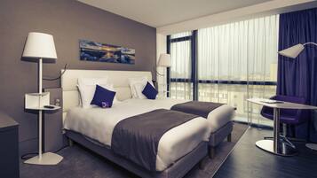 A hotel room with a bed, two bedside tables, a desk with a chair, and a large window with curtains.