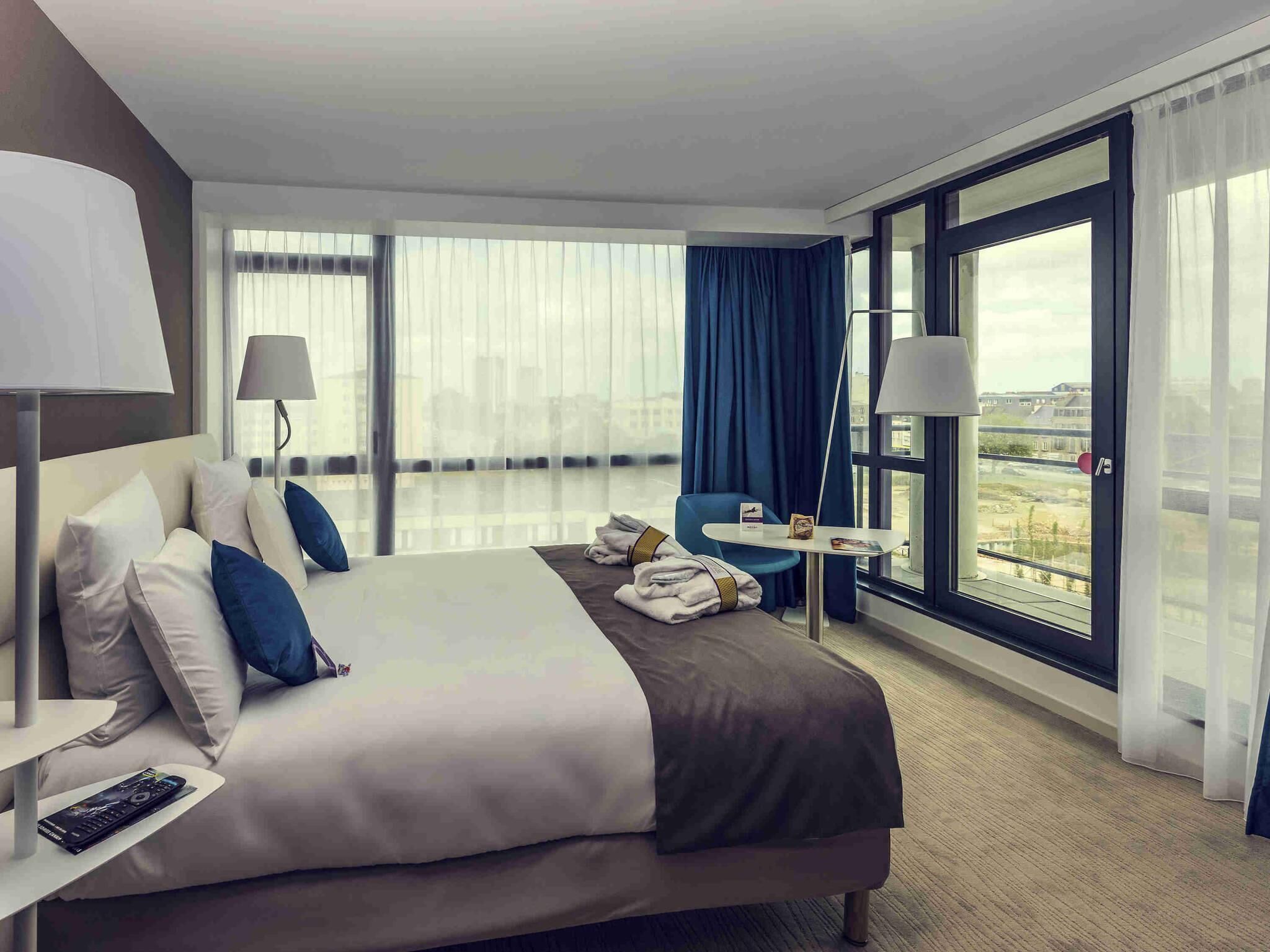 A hotel room with a large bed, a small table, a chair, and a view of the city.