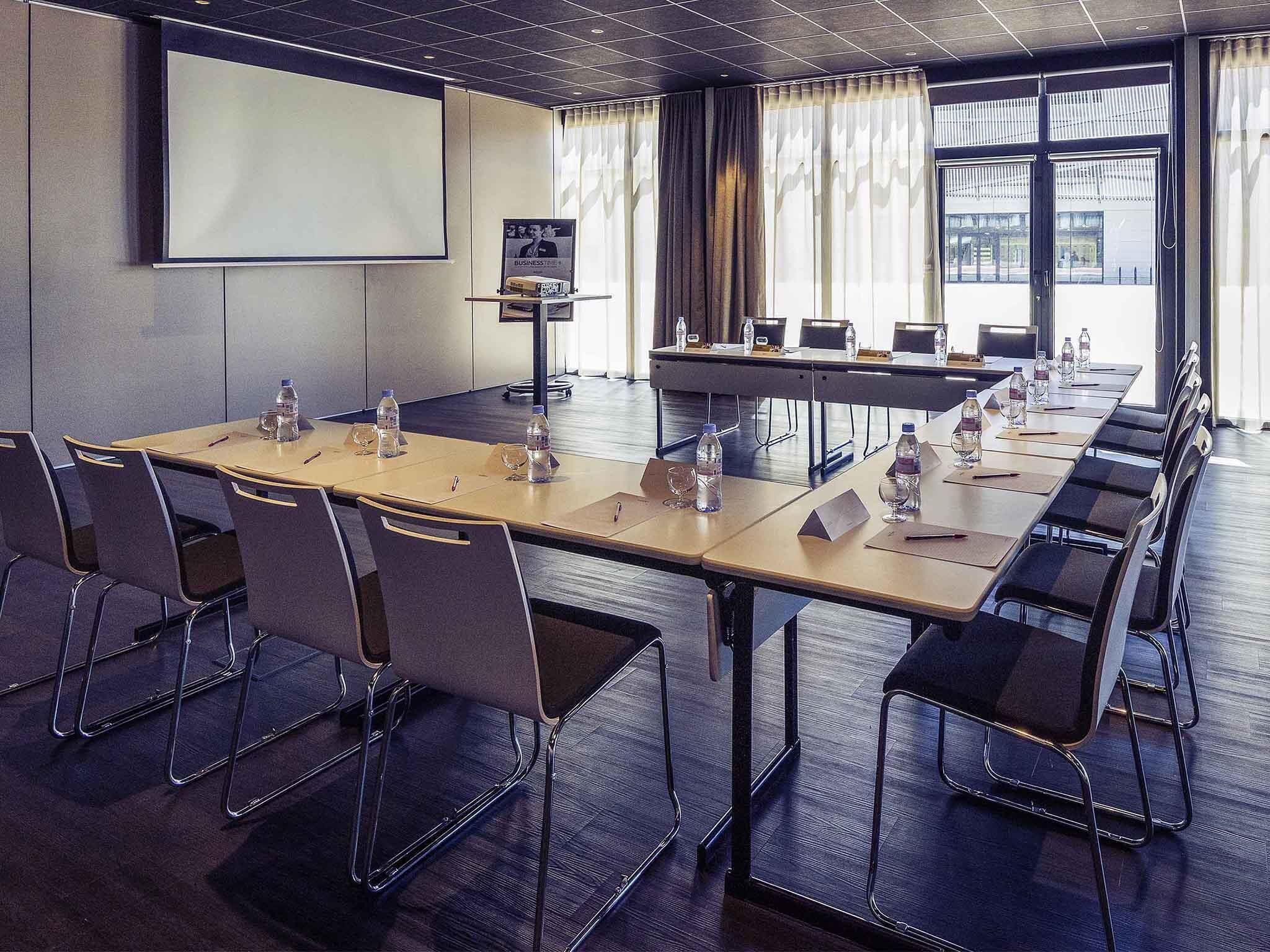 Una sala de conferencias con una larga mesa, sillas, una pantalla de proyector y un amplio ventanal con cortinas.