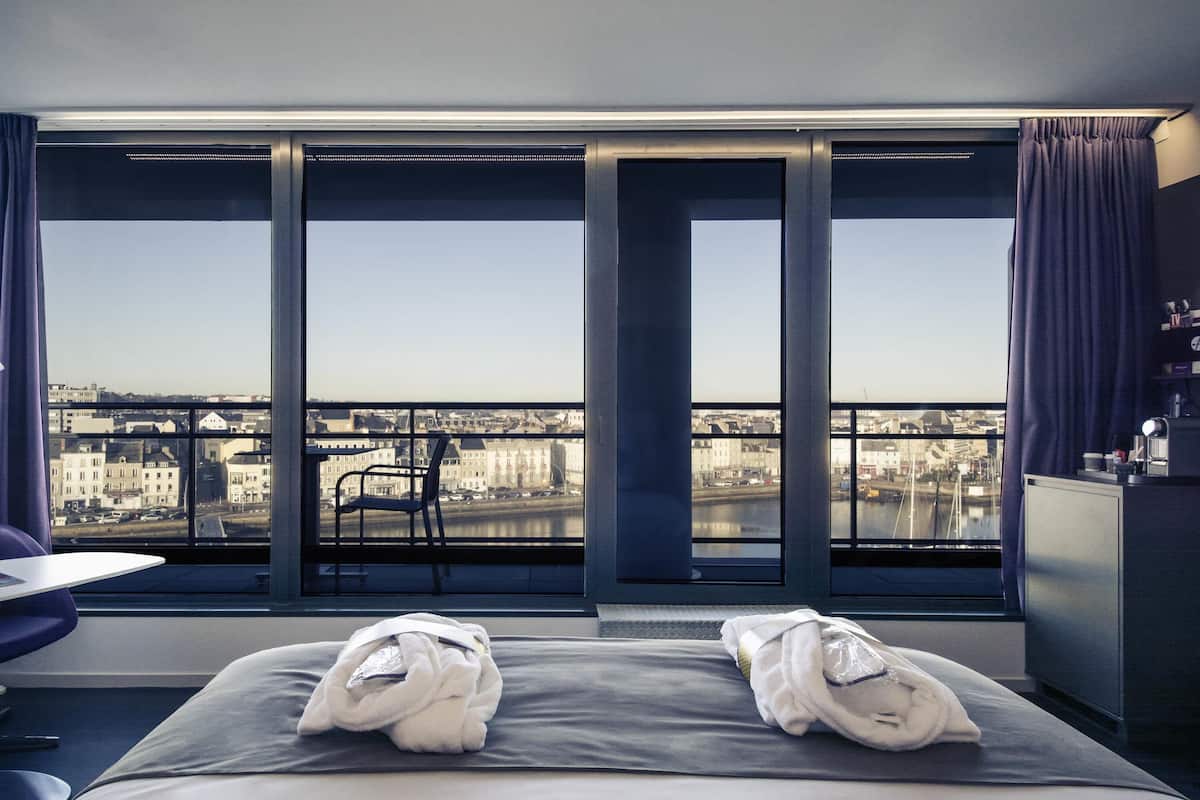 A hotel room with a large window, a bed with towels, a chair, and a desk.
