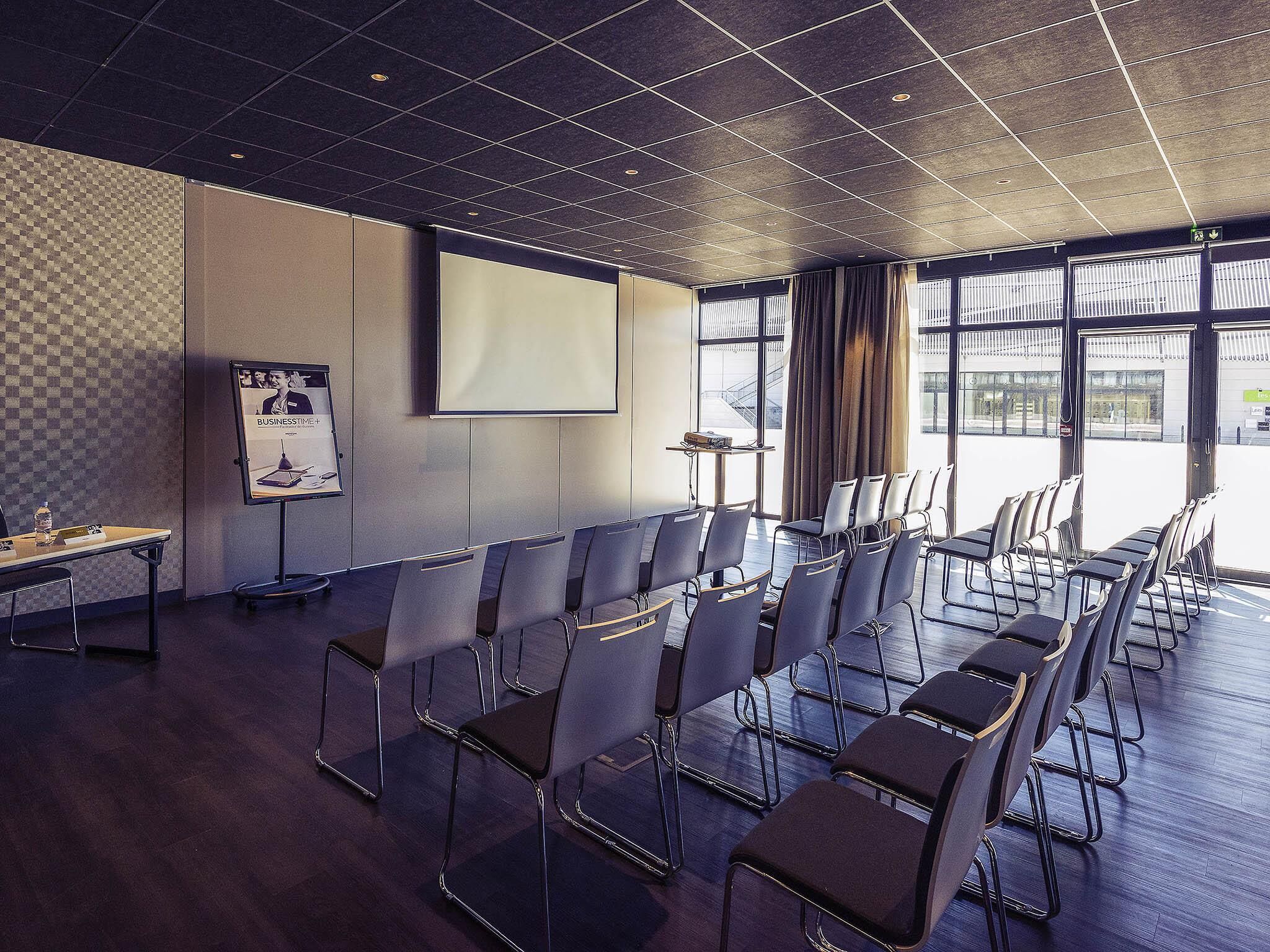 Un salón de conferencias con una pantalla grande, hileras de sillas, un proyector y una pizarra para presentaciones.