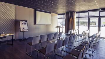 Un salón de conferencias con una pantalla grande, hileras de sillas, un proyector y una pizarra para presentaciones.