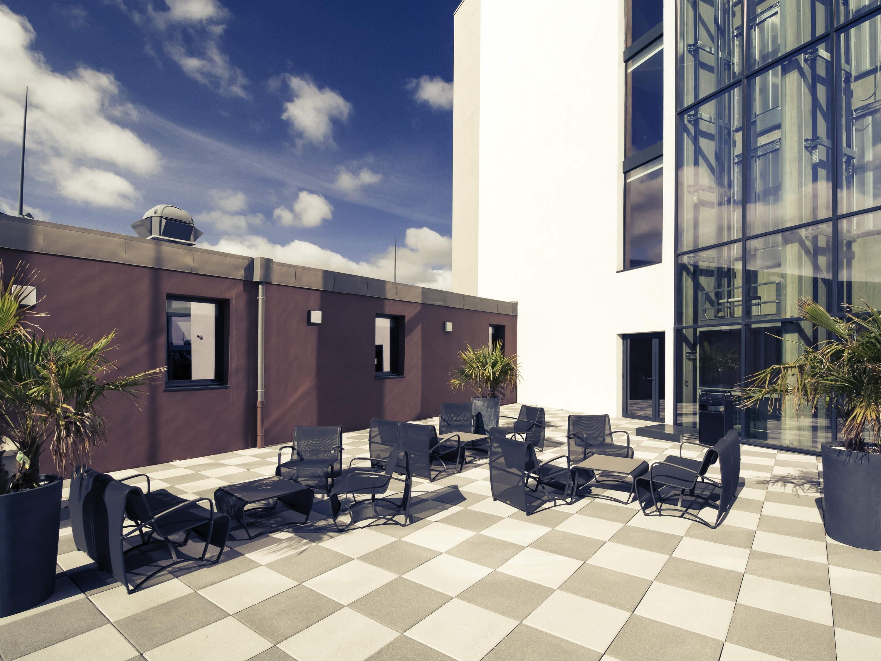 Une terrasse sur le toit, aménagée avec des espaces de détente en plein air, des plantes en pot et un bâtiment moderne doté de grandes fenêtres.