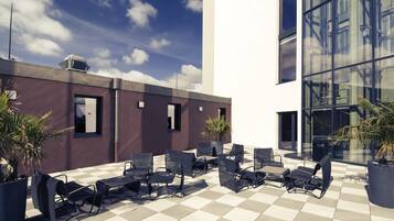 Une terrasse sur le toit, aménagée avec des espaces de détente en plein air, des plantes en pot et un bâtiment moderne doté de grandes fenêtres.