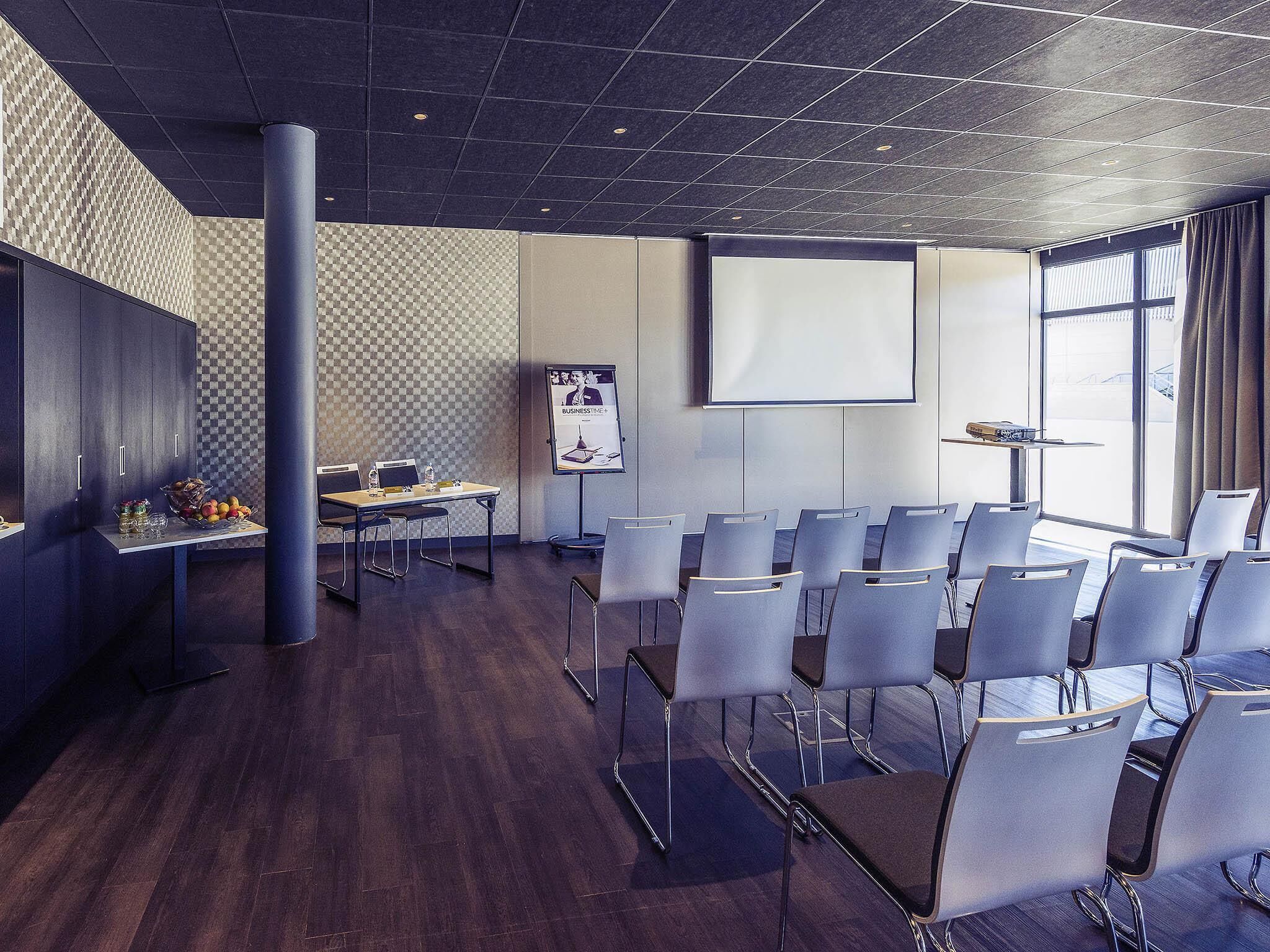 Uma sala de conferências com tela de projeção, quadro branco e uma mesa com frutas e garrafas de água.