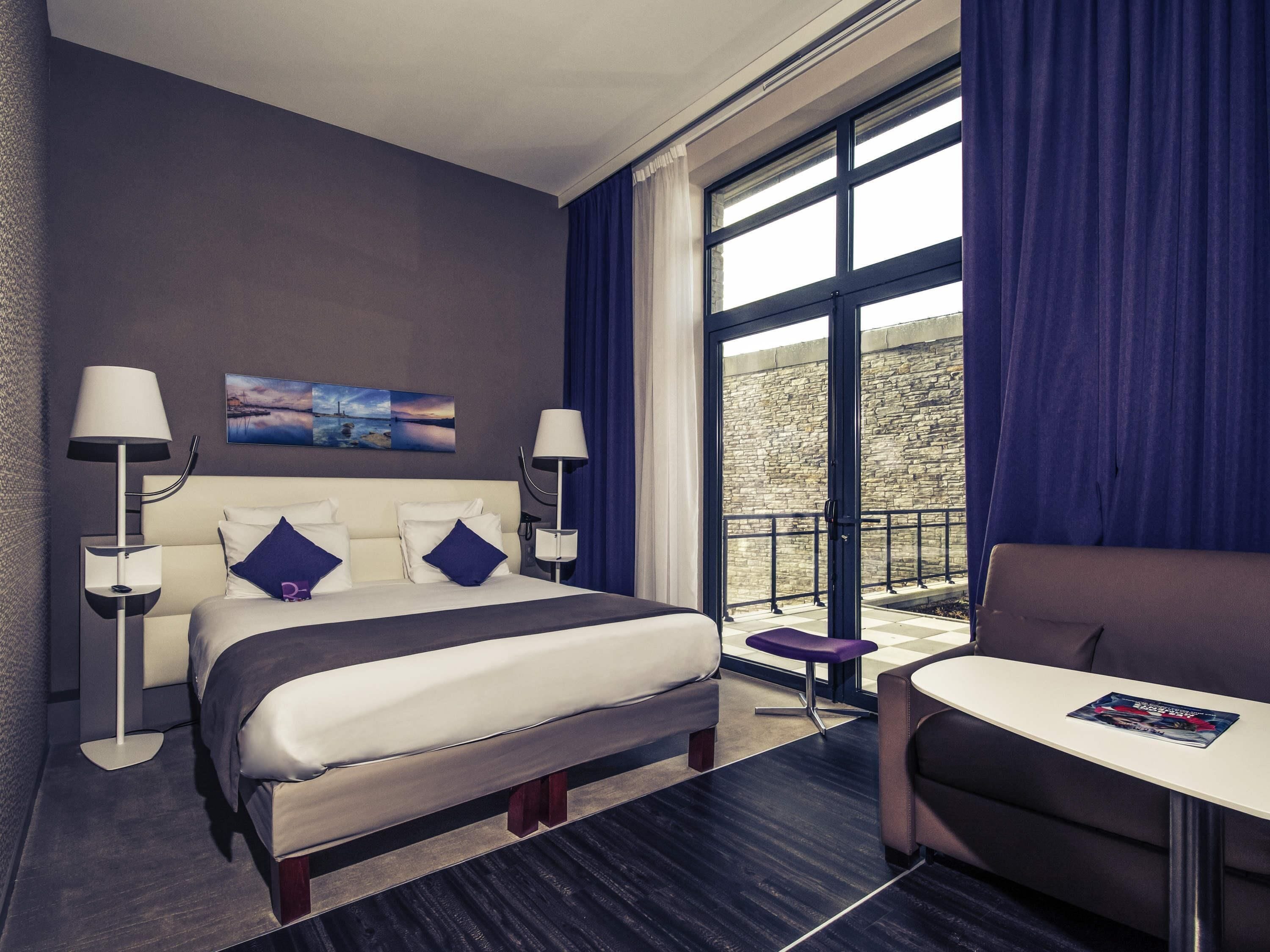 Een hotelkamer met een bed, twee bedlampen, een kleine tafel, een bank en een groot raam met gordijnen.