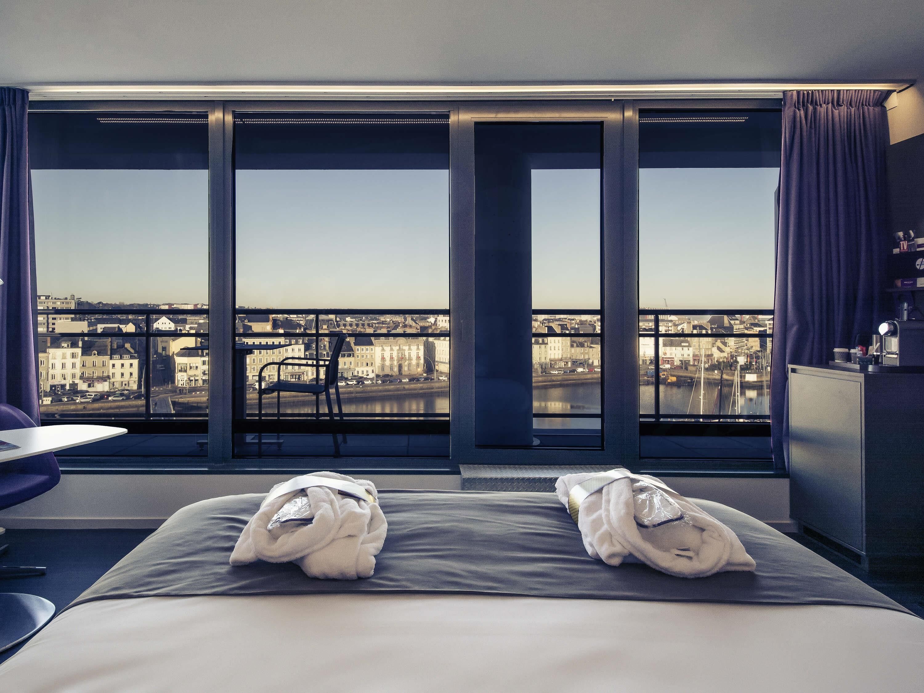 A hotel room with a large window, a bed with towels, a chair, and a desk.