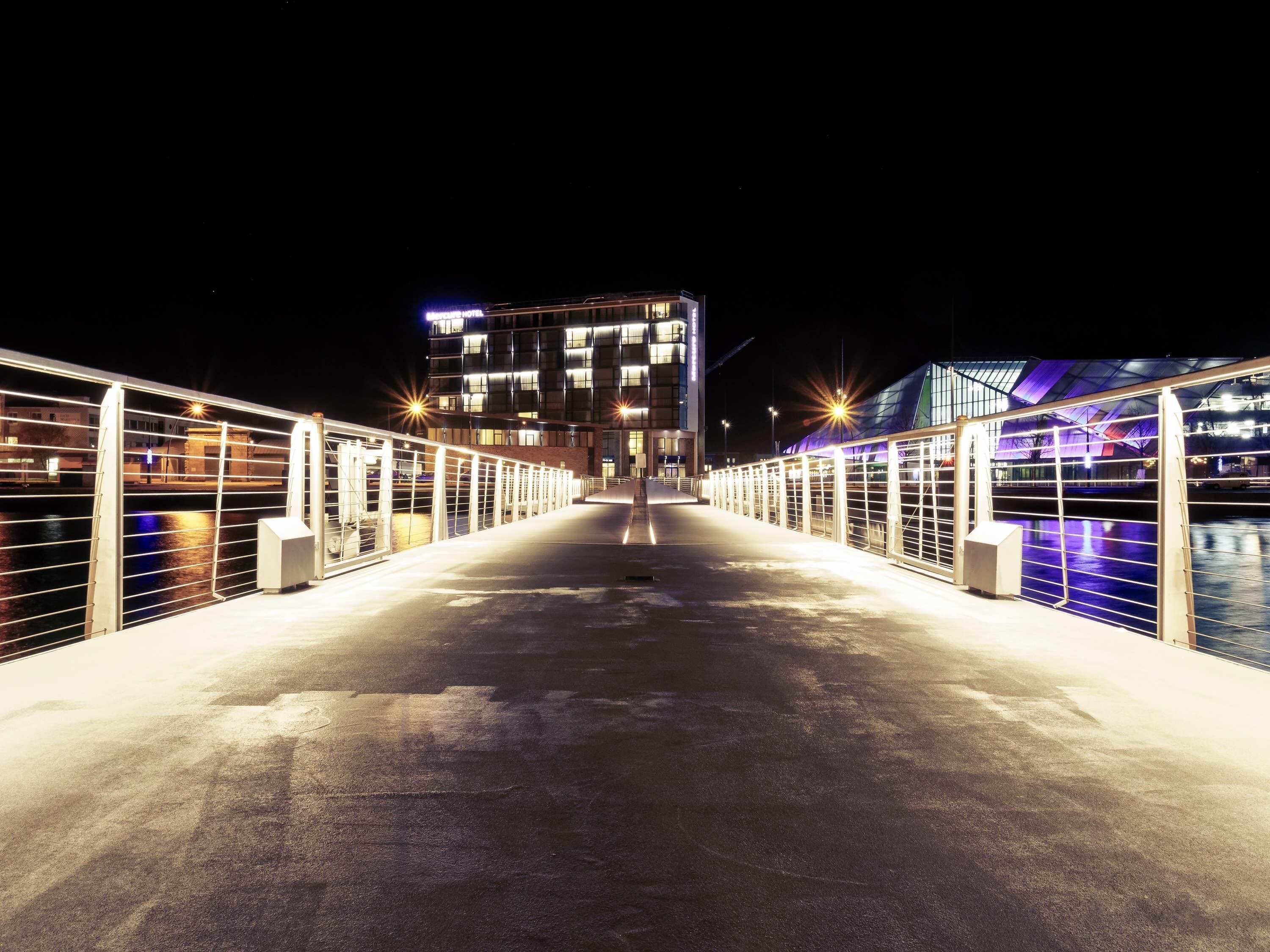 Une vue nocturne d’un pont menant à un bâtiment moderne à plusieurs étages, doté de balcons et relié à un autre bâtiment par une passerelle avec des garde-corps.