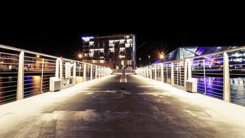 Une vue nocturne d’un pont menant à un bâtiment moderne à plusieurs étages, doté de balcons et relié à un autre bâtiment par une passerelle avec des garde-corps.