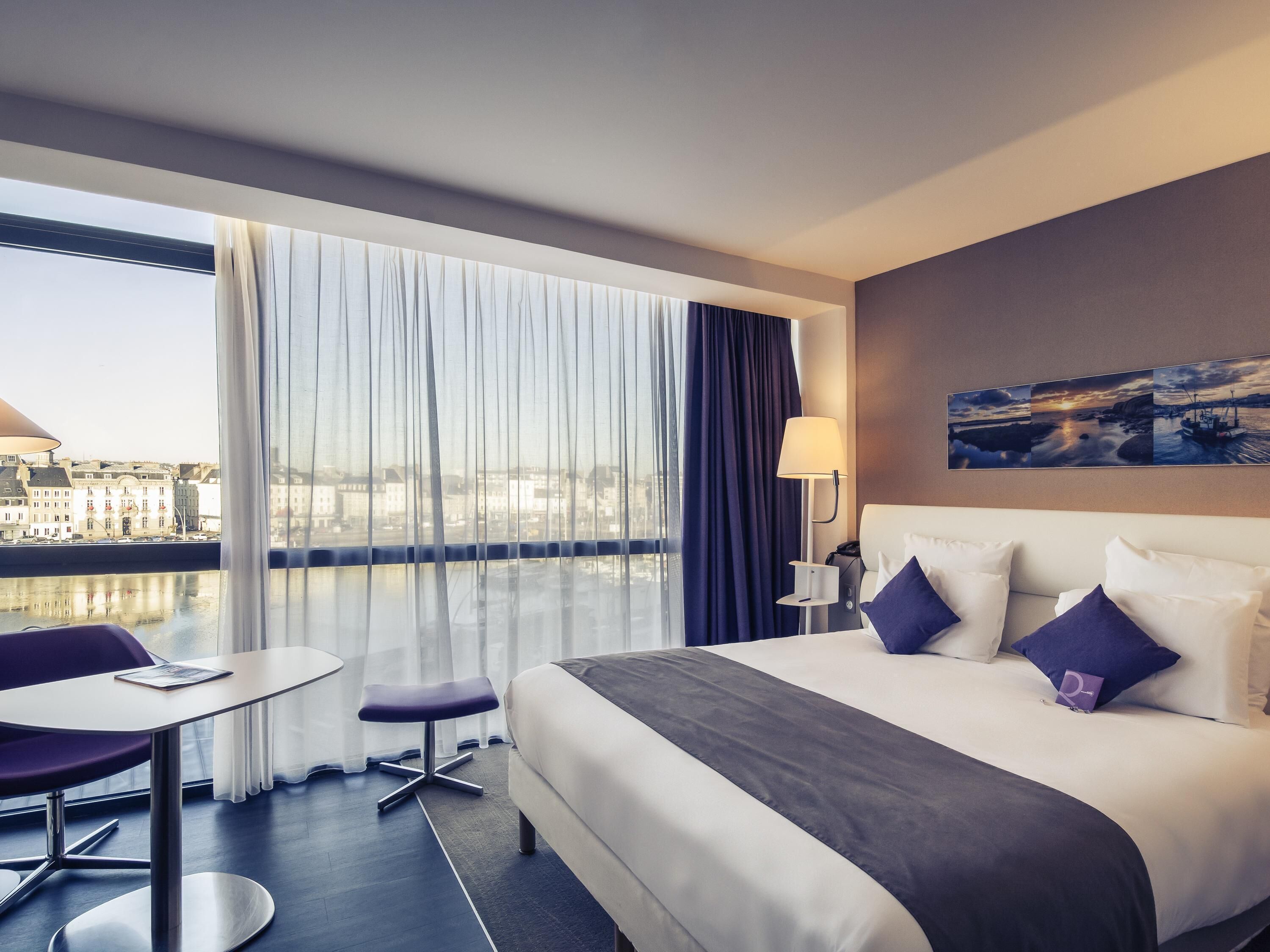 Habitación de hotel con cama, escritorio, silla, lámpara y vista a un paisaje urbano a través de amplias ventanas.