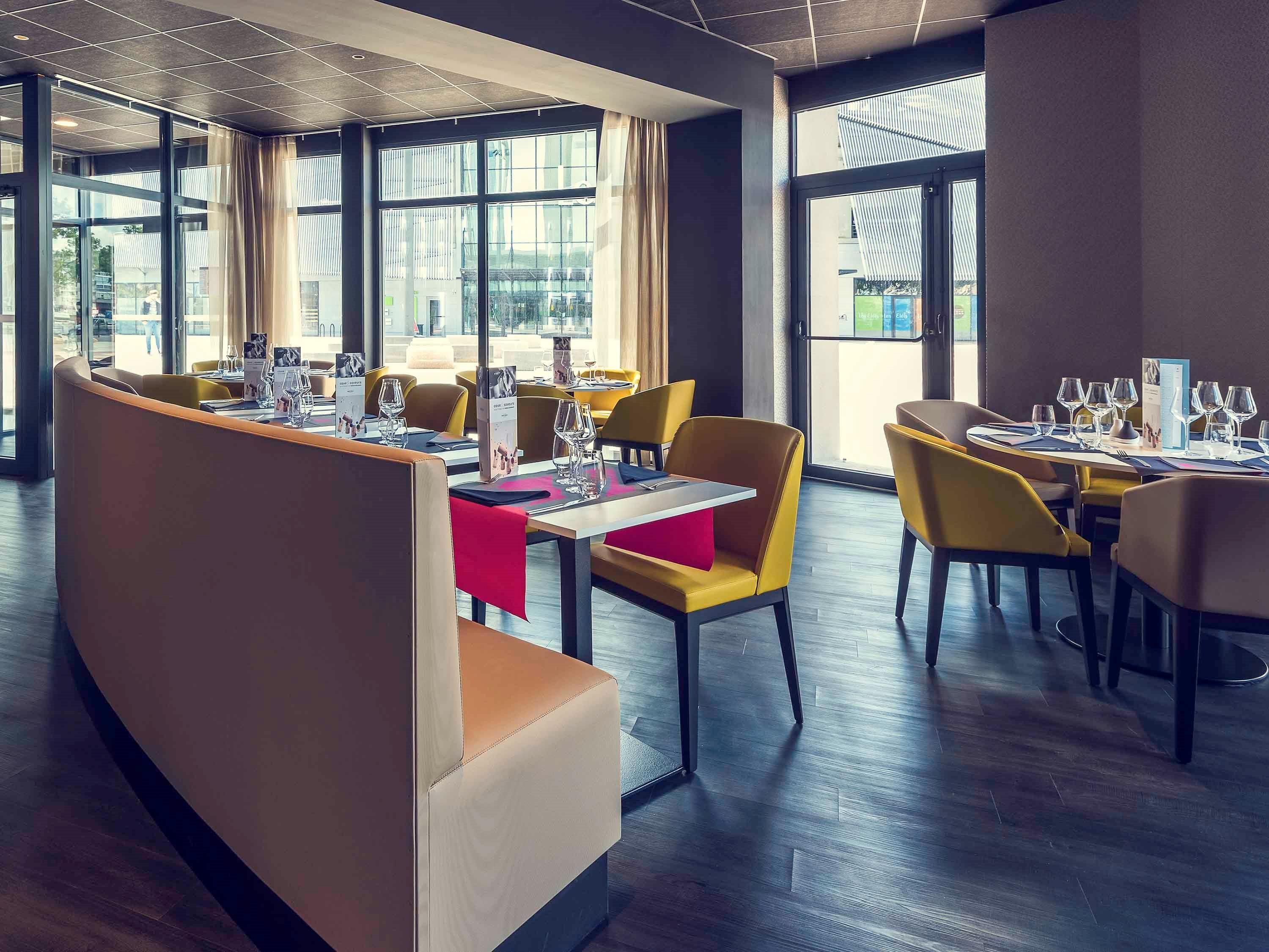 A modern restaurant with high ceilings, large windows, and a mix of yellow and brown seating. Tables are set with wine glasses and menus.