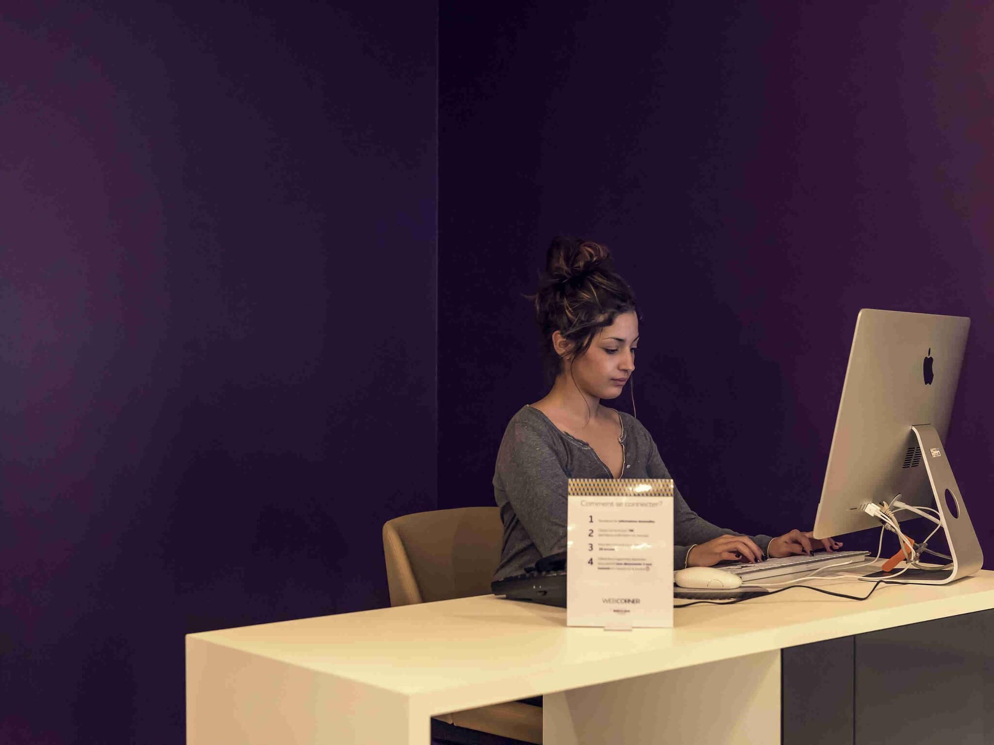 Une personne utilise un MacBook sur un bureau, et une affiche sur laquelle on peut lire « Comment se connecter ? » est visible dans la pièce, dont les murs sont de couleur violette.