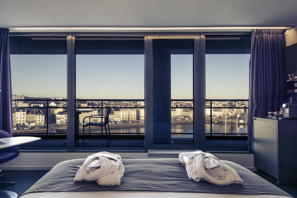 Une chambre d’hôtel avec une grande fenêtre, un lit avec des serviettes, une chaise et un bureau.