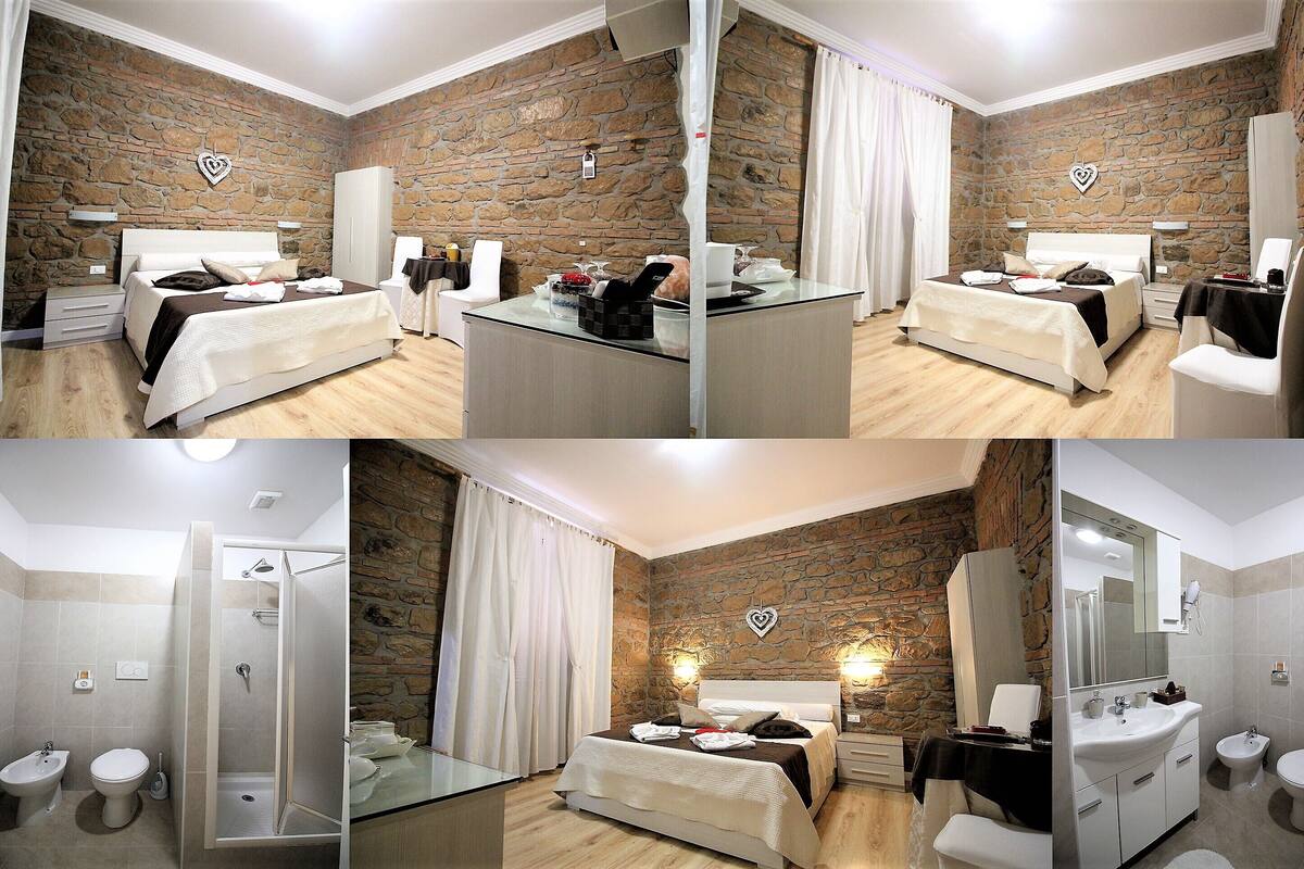 A hotel room with a stone wall, a bed with white linens, a bedside table with a lamp, a shower area, and a bathroom with a sink and toilet.