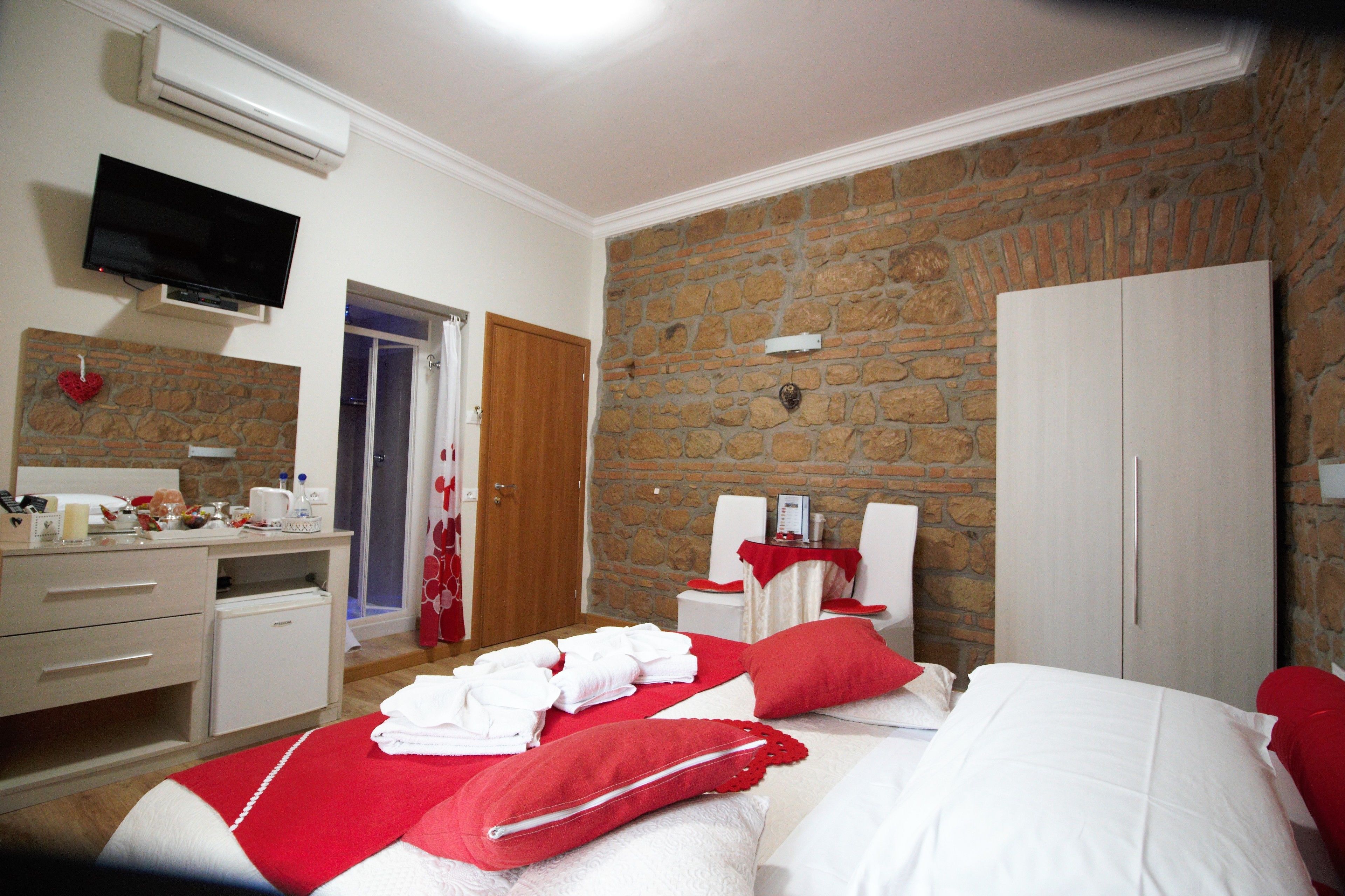 Une chambre d’hôtel avec un mur en pierre, un lit avec une literie rouge et blanche, un petit bureau avec un miroir, une télévision et une armoire.