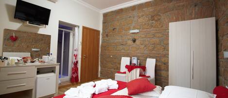 Une chambre d’hôtel avec un mur en pierre, un lit avec une literie rouge et blanche, un petit bureau avec un miroir, une télévision et une armoire.