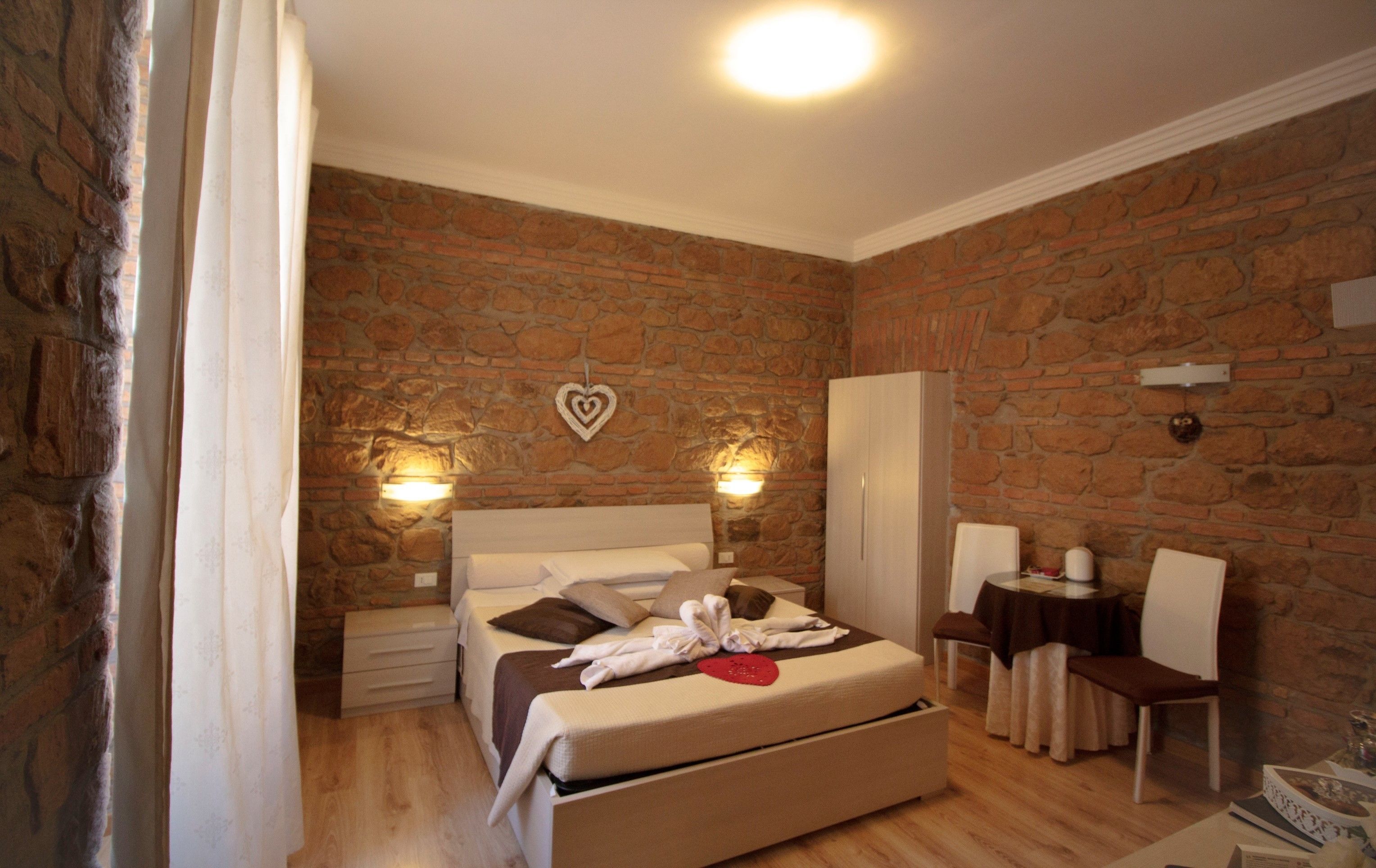 A bedroom with a stone wall, a bed with a headboard, bedside tables, a wardrobe, a desk with a chair, and a small table with a lamp.