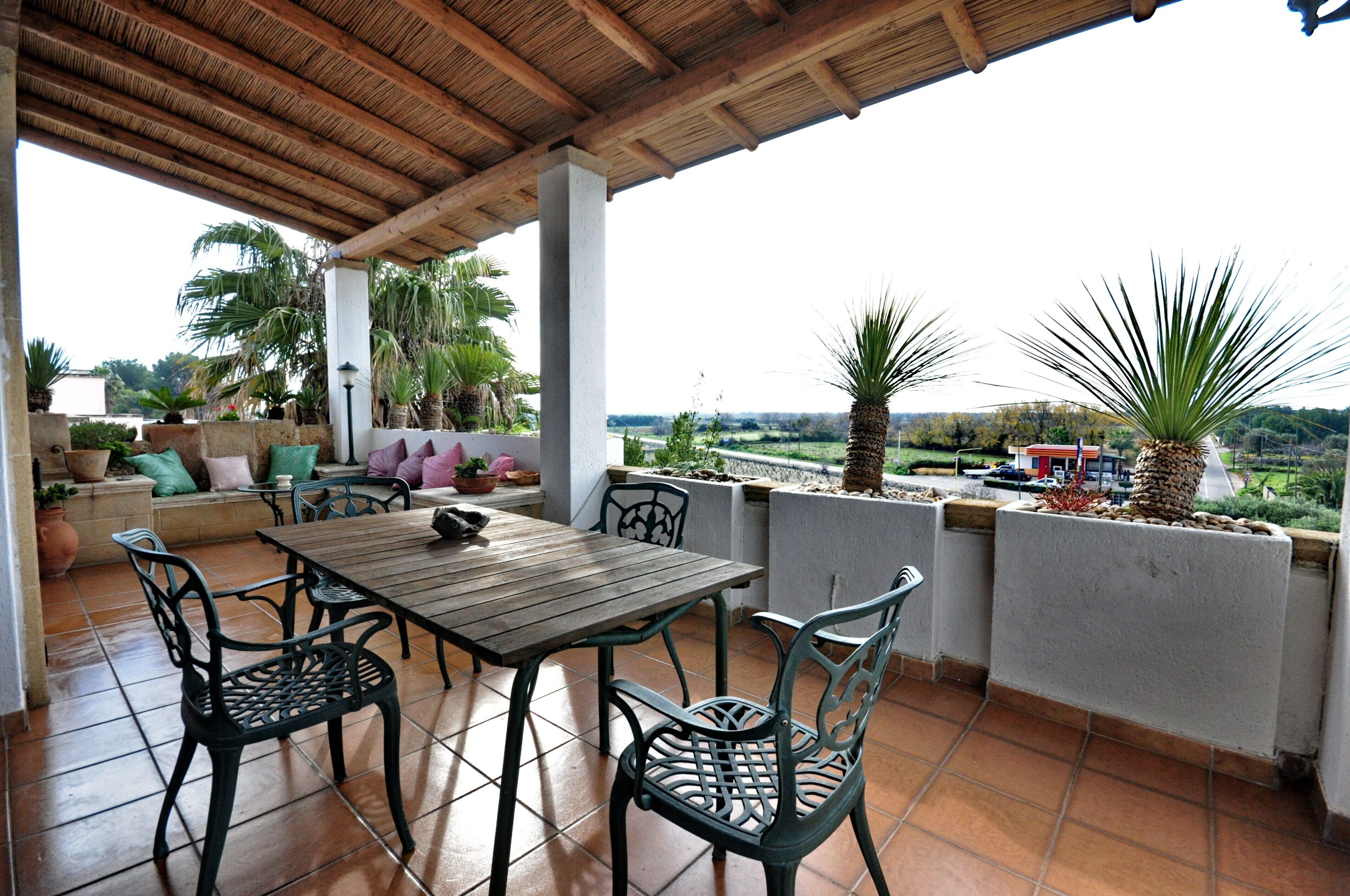 A covered patio with a wooden table and chairs, surrounded by potted plants and a view of the countryside.