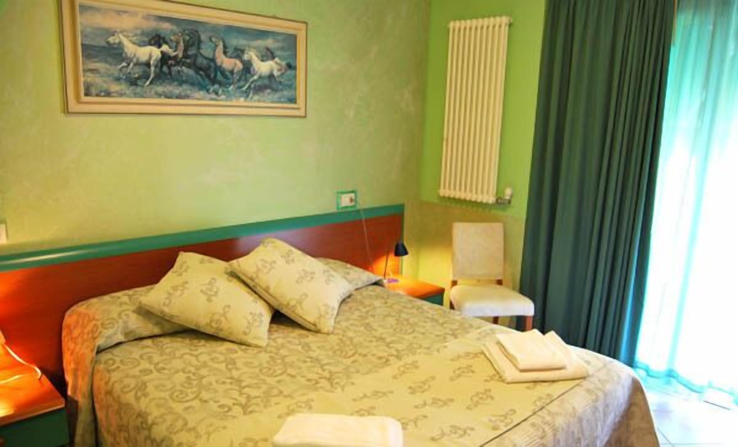 A hotel room with a bed, pillows, a chair, a painting on the wall, and a window with curtains.