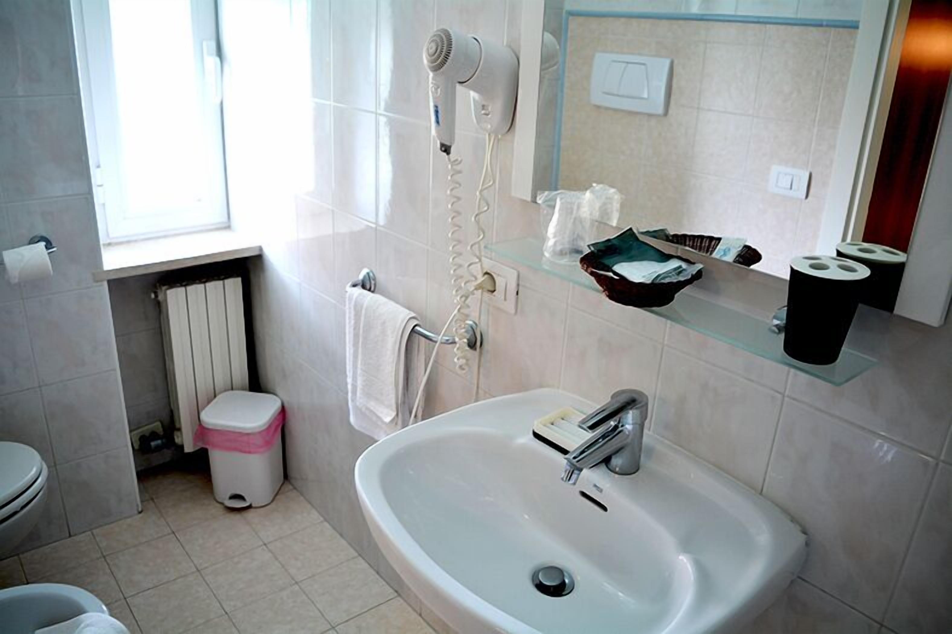 A bathroom with a sink, a hairdryer, a towel rack, a glass shelf with toiletries, and a window.