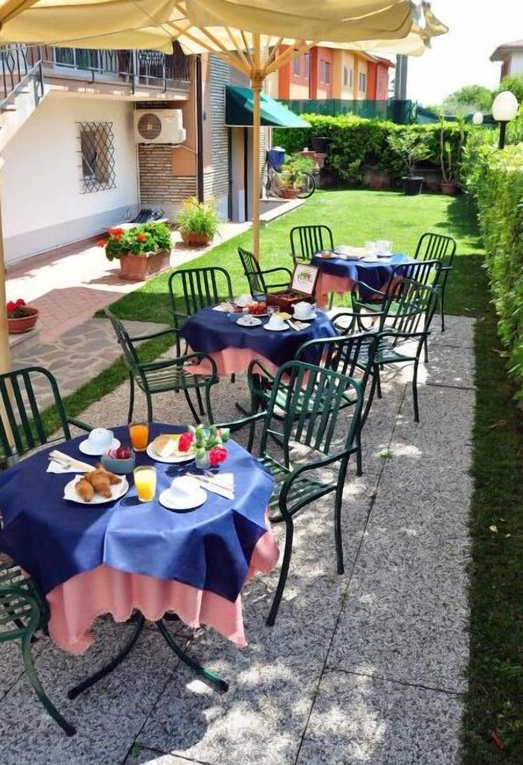 Un espace de restauration en plein air avec des tables dressées pour le petit-déjeuner, entouré de verdure et avec un bâtiment en arrière-plan.