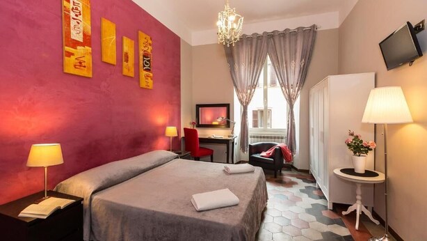 A hotel room with a bed, bedside table, lamp, TV, desk, chair, and a view of the outside. - Claudia House Roma Termini (Rome)