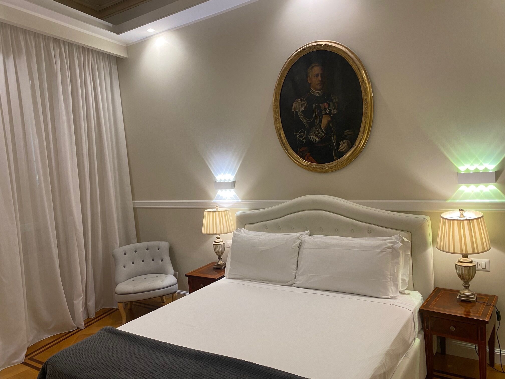 Una camera d'albergo con un letto grande, due abat-jour, una sedia, un quadro appeso al muro e una finestra con tende.