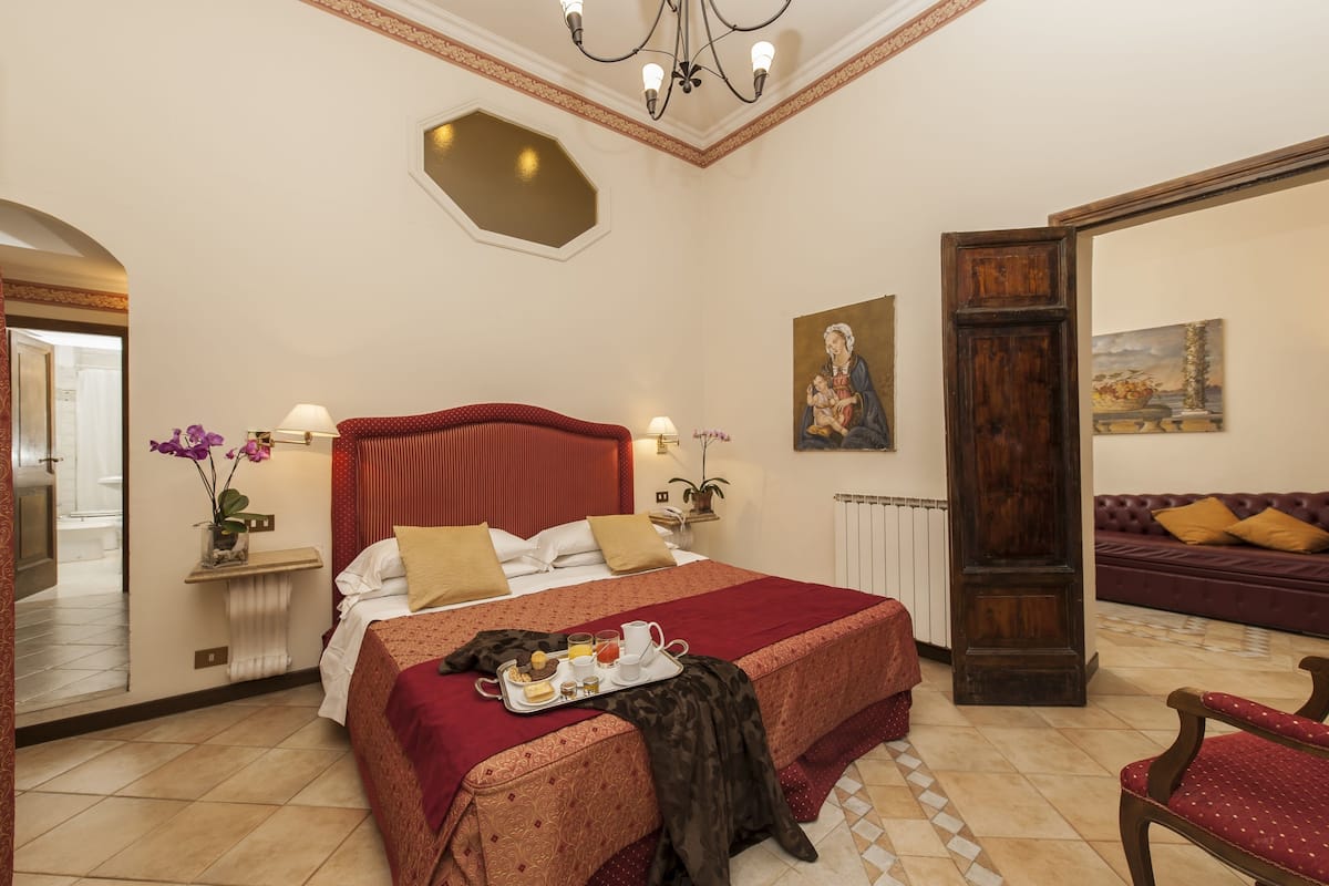 Ein Hotelzimmer mit einem großen Bett, einer Sitzecke mit einer Couch, einem Esstisch mit Frühstück und Blick auf den Flur.
