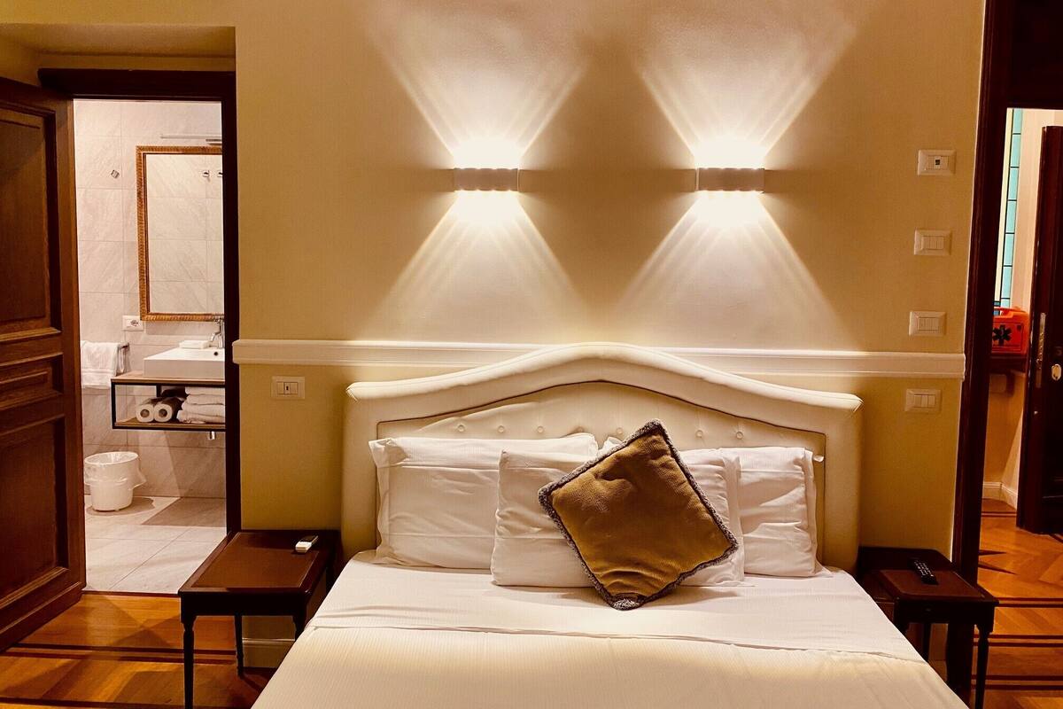 Ein Hotelzimmer mit einem großen Bett, einem Nachttisch, einem durch eine offene Tür einsehbaren Badezimmer und Wandleuchten.