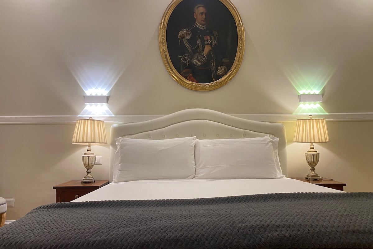 Ein ordentlich bezogenes Bett mit weißen Leinen und grauer Decke, flankiert von zwei Nachttischen mit Lampen, und ein gerahmtes Porträt darüber.