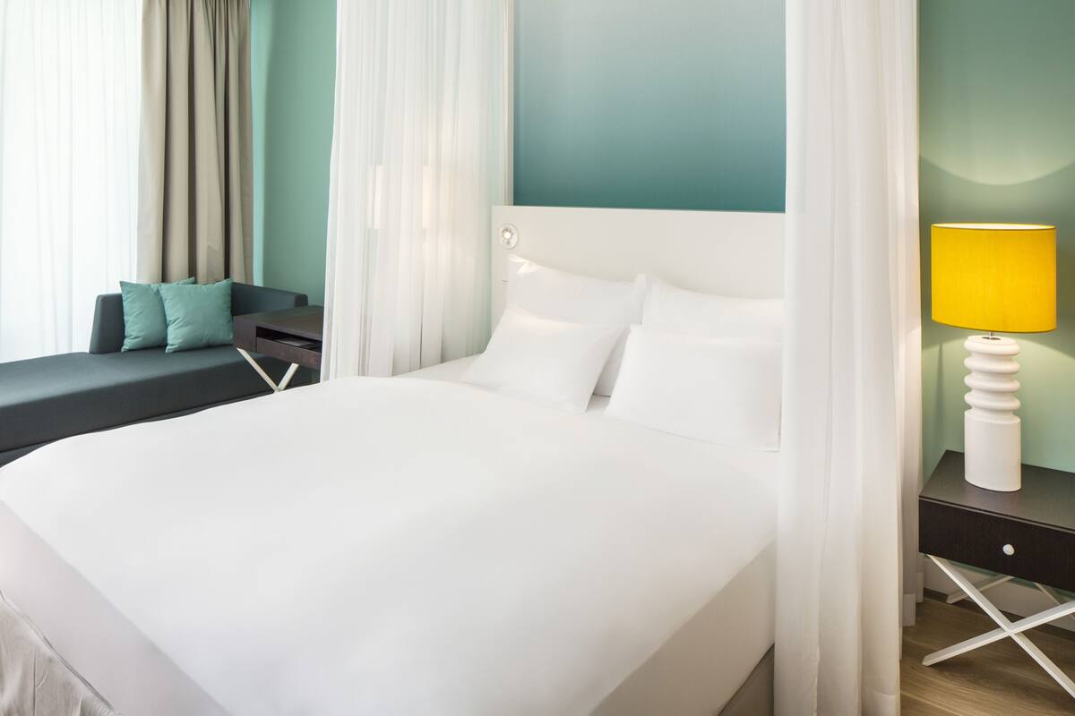 A modern hotel room with a large bed, a bedside table with a lamp, a sofa with cushions, and sheer curtains.