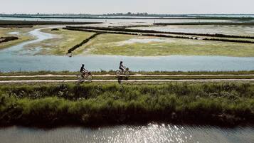 Una persona pedaleando en bicicleta a lo largo de una vía fluvial con riberas cubiertas de hierba y un paisaje urbano a lo lejos.