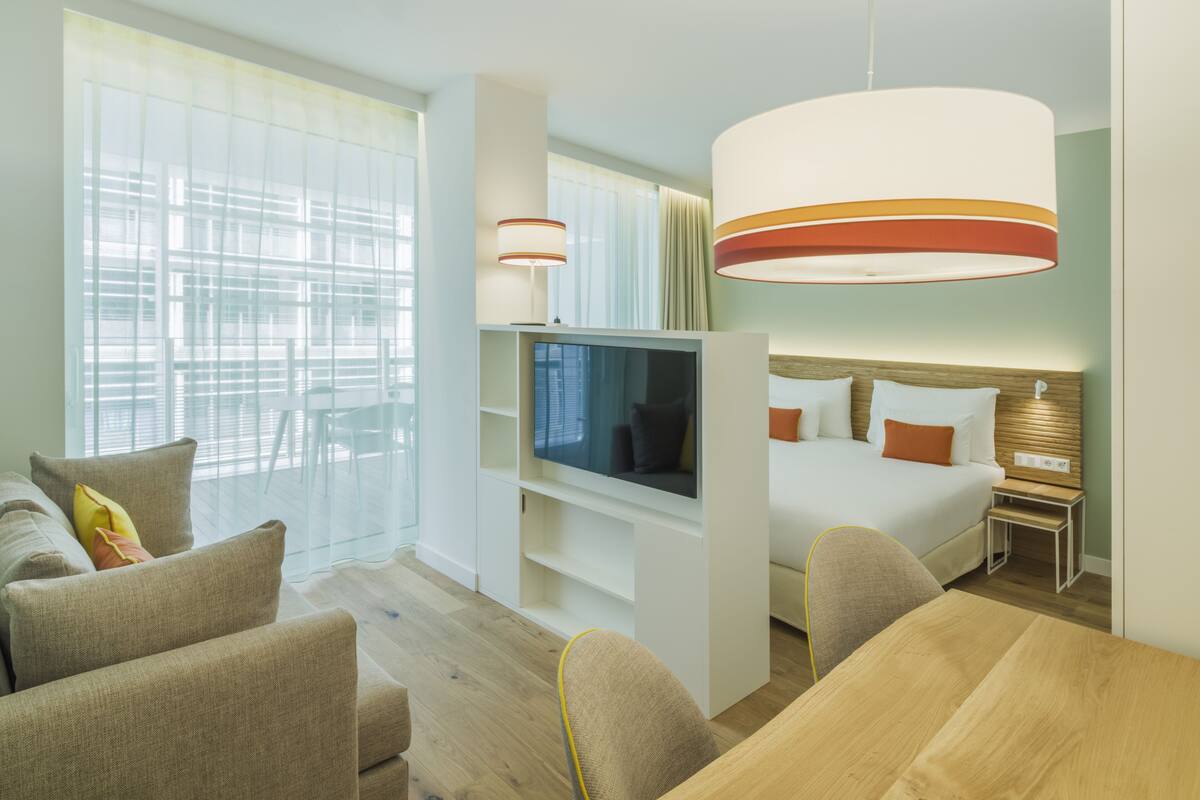 A modern hotel room with a large bed, a TV, a sofa, and a dining table.