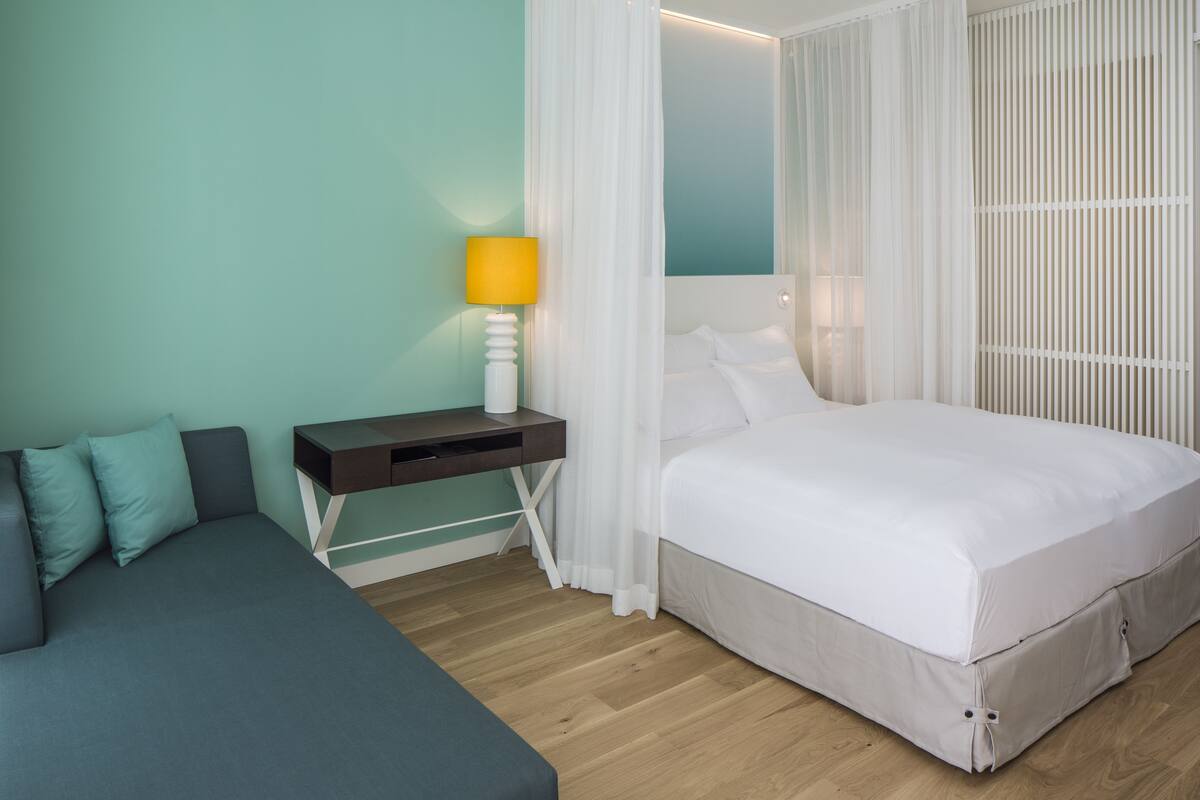 A bedroom with a bed, a bedside table with a lamp, a nightstand, and a sofa.