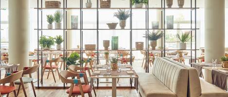 A modern dining area with wooden flooring, multiple tables and chairs, a sofa, and a large window with potted plants.