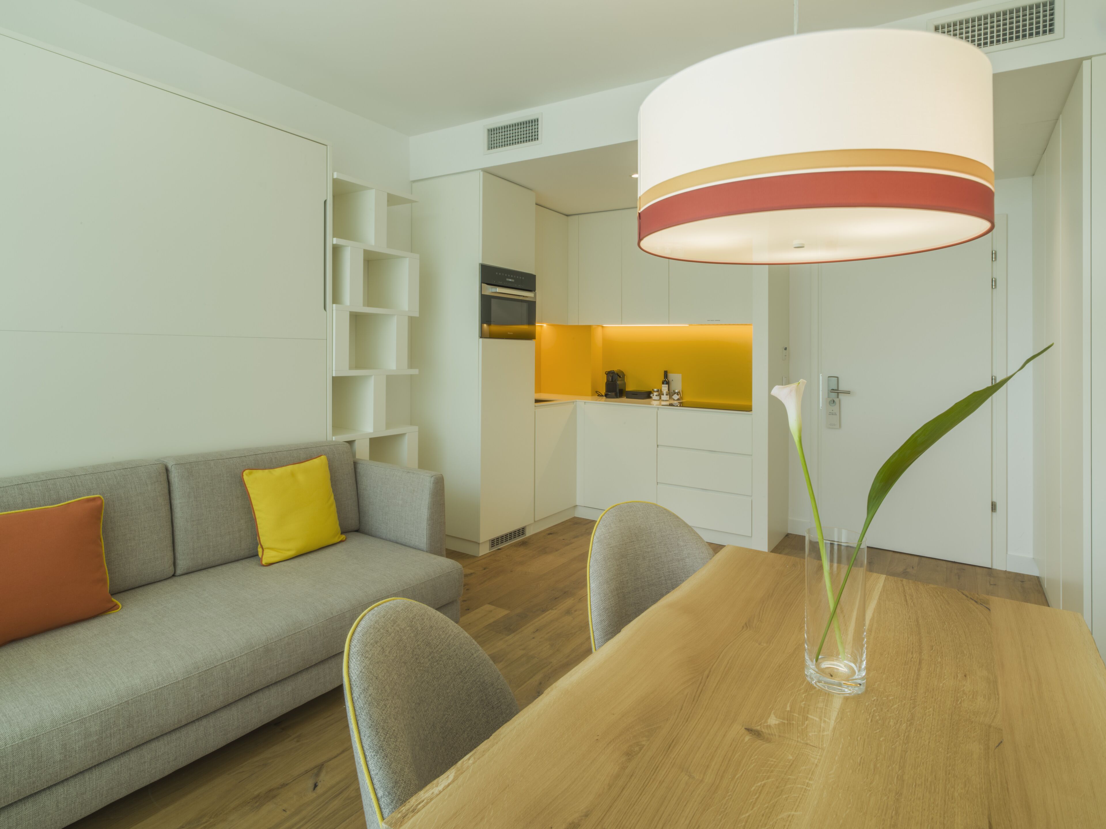 Un salon moderne avec une table à manger en bois, un canapé gris agrémenté de coussins orange et un vase transparent contenant une seule plante.