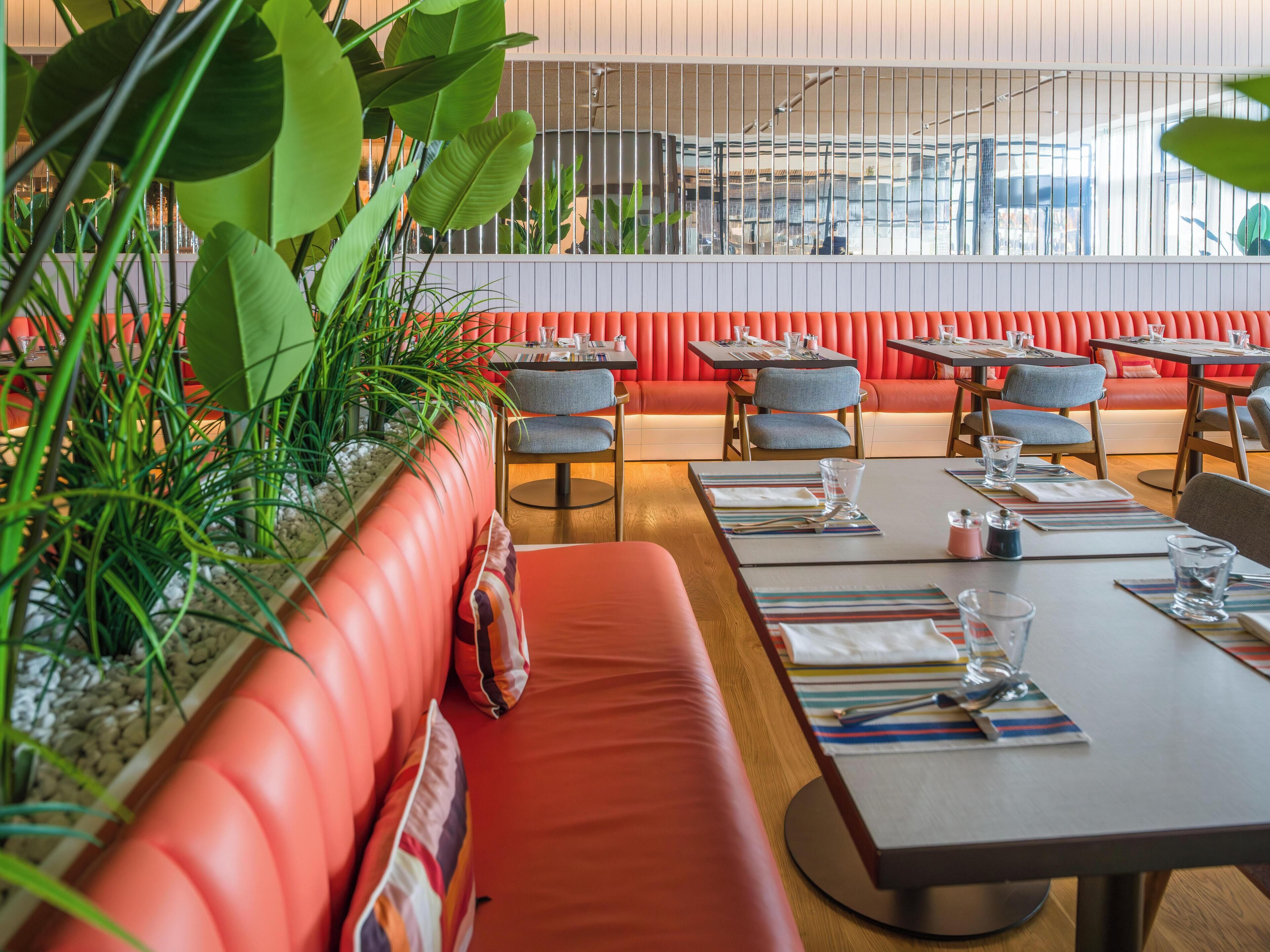 Un restaurant avec des banquettes et des tables recouvertes d’un tissu rouge, et offrant une vue sur l’extérieur à travers une fenêtre dotée de stores verticaux.